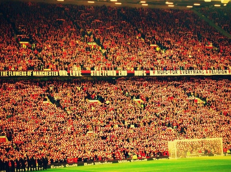 Stretford End chật cứng khán giả, biển người đứng san sát, vẫy cờ và khăn quàng.